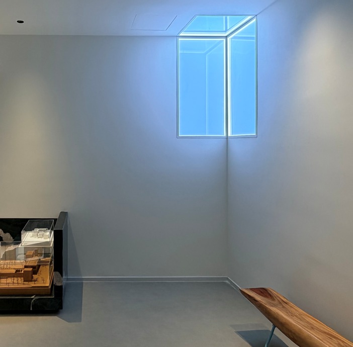 Blue-lit, window-like panel set high in a white wall above a scale model and timber bench